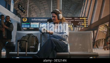 La femme retire les gros écouteurs, saisit le sac à dos et la valise, et se dirige vers la porte de départ. Concept de transition de l'attente au voyage, l'excitation de commencer le voyage dans l'aéroport moderne. Banque D'Images