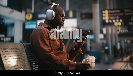 Black Man s'assoit confortablement dans la zone d'attente de l'aéroport, portant des écouteurs élégants argentés au-dessus de l'oreille. Gars appréciant Hamburger, prenant des bouchées pendant Lost in Music. Concept de style de vie du passager moderne. Banque D'Images