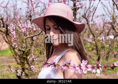 Femme portant un chapeau de soleil rose et un top blanc, debout dans un verger de pêchers en fleurs ensoleillées, entourée de belles fleurs roses sur les branches pendant le sprin Banque D'Images