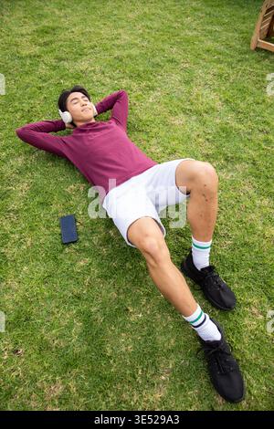 Homme asiatique adulte couché sur la pelouse verte portant des écouteurs et des shorts avec smartphone à proximité Banque D'Images