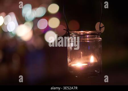 Une photographie rapprochée d'une bougie allumée brûlant à l'intérieur d'une lanterne en verre transparent avec une poignée en fil noir, suspendue sur un fond sombre de nuit. Banque D'Images