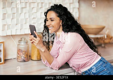 Femme sourit tout en utilisant smartphone dans la cuisine pendant la journée Banque D'Images
