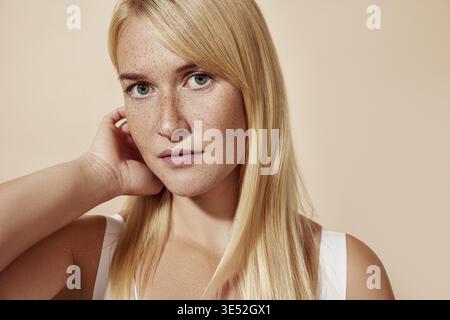 Jeune belle femme blonde avec des taches de rousseur regardant la caméra et ajustant ses cheveux Banque D'Images