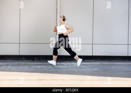 Vue latérale d'une femme grande taille portant des écouteurs près d'un mur. Femme courbée dans des vêtements de sport jogging à l'extérieur Banque D'Images