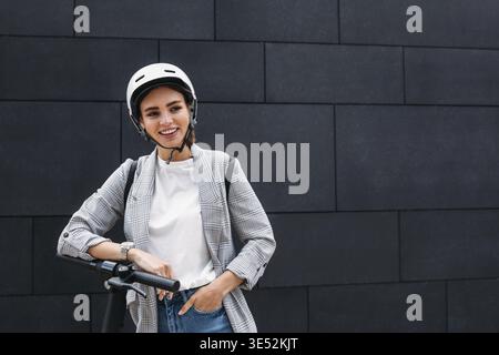 Portrait d'une femme d'affaires souriante penchée sur un guidon de scooter électrique. Jeune femme gaie dans un casque de vélo au mur noir qui regarde loin Banque D'Images