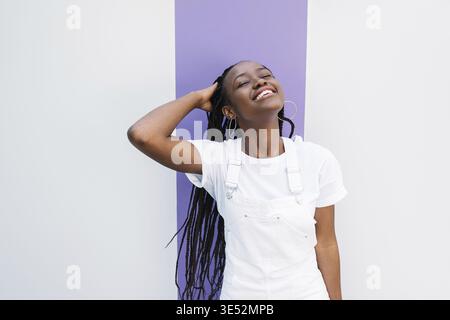 Jeune femme souriante dans les décontractés ajustement des cheveux avec les yeux fermés contre le mur blanc avec une bande mauve Banque D'Images
