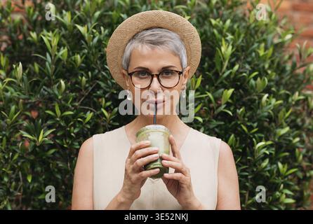 Femme mature élégante avec les cheveux gris buvant du matcha latte à l'extérieur Banque D'Images