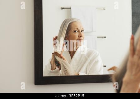 Femme âgée regardant le miroir tout en peignant ses cheveux longs dans la salle de bains Banque D'Images