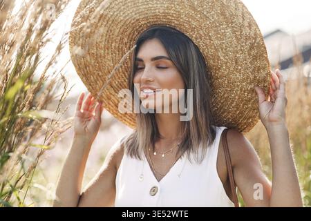 Belle femme blonde touchant son chapeau de paille avec les yeux fermés Banque D'Images