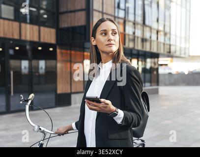 Jeune femme d'affaires confiante avec téléphone portable et vélo debout à l'extérieur portant des vêtements formels Banque D'Images
