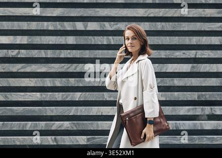Vue latérale de femme d'affaires élégante d'âge moyen en manteau. Femme d'âge moyen parlant au téléphone portable tout en marchant Banque D'Images