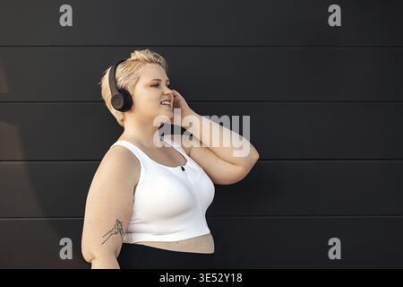 Vue latérale d'une femme souriante dans des vêtements de fitness écoutant de la musique à un mur noir Banque D'Images