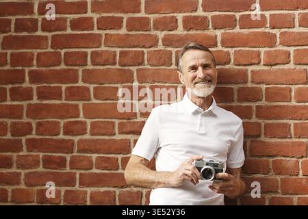 Homme mature souriant regardant la caméra. Touriste avec caméra debout au mur de briques Banque D'Images