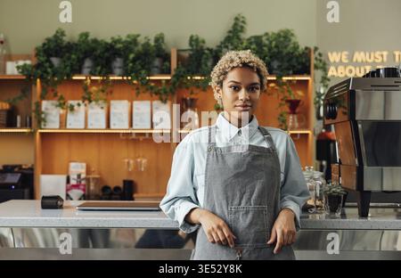 Barista confiante s'appuyant sur un comptoir regardant la caméra. Jeune femme dans un tablier travaillant comme barista debout dans un café Banque D'Images