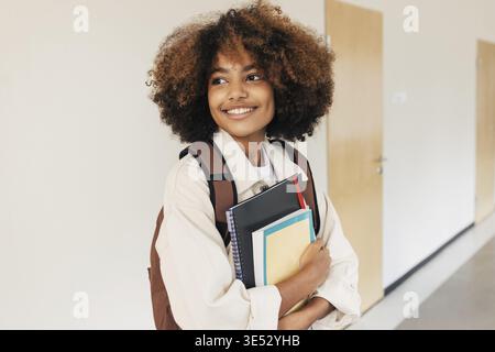 Étudiant souriant avec les cheveux bouclés debout dans le couloir et regardant loin. Fille avec des manuels et un ordinateur portable à l'école Banque D'Images