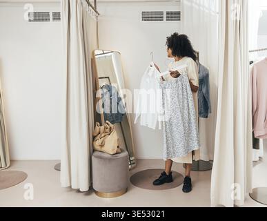 Jeune femme choisissant des vêtements tout en se tenant dans les cabines d'essayage dans une boutique Banque D'Images