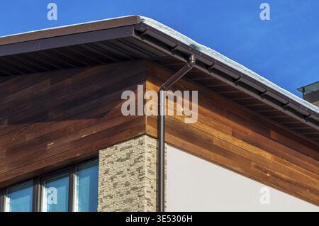 Gros plan d'un coin de bâtiment avec des boiseries, des accents de pierre, des gouttières brunes contre un ciel bleu clair et un soupçon de neige sur le toit Banque D'Images