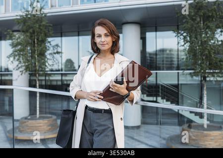 Femme d'âge moyen confiante tenant un dossier debout contre la construction d'entreprise Banque D'Images