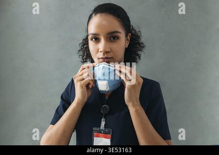 Infirmière afro-américaine tenant un masque de protection regardant la caméra au mur Banque D'Images