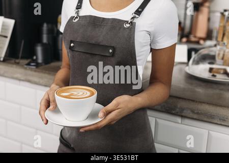 Photo recadrée d'un barista méconnaissable tenant une tasse de café. Serveuse dans un tablier avec un cappuccino fraîchement préparé Banque D'Images