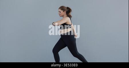 Jogging féminin de grande taille sur un mur gris. Femme avec un corps grande taille courir à l'extérieur et regarder sa smartwatch Banque D'Images