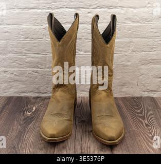 Une paire de bottes de cow-boy occidental en cuir brun vieilli se tient sur un plancher de bois sombre contre un fond de mur de briques blanches. Banque D'Images
