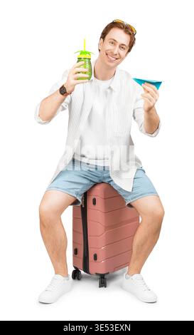 Heureux jeune homme prêt pour les vacances d'été assis sur une valise rose, tenant une boisson tropicale et un avion de papier bleu, isolé sur une grotte blanche Banque D'Images