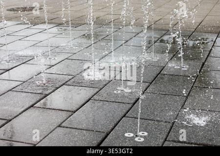 Des jets d'eau projettent vers le haut depuis le sol dans une fontaine moderne, créant des ruisseaux ludiques sur des carreaux de pierre gris. La conception géométrique propre ajoute une réf Banque D'Images