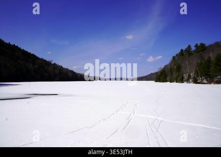 Une vaste étendue enneigée de lac Philippe gelé est encadrée par des collines densément boisées sous un ciel bleu vif dans le parc de la Gatineau, au Québec Banque D'Images