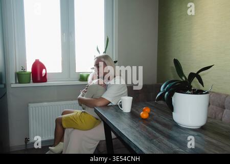 Une mère et son fils prennent le petit déjeuner dans une cuisine moderne le matin. L'enfant heureux et affamé mange. Une femme endormie, sans maquillage, d'apparence naturelle, Banque D'Images