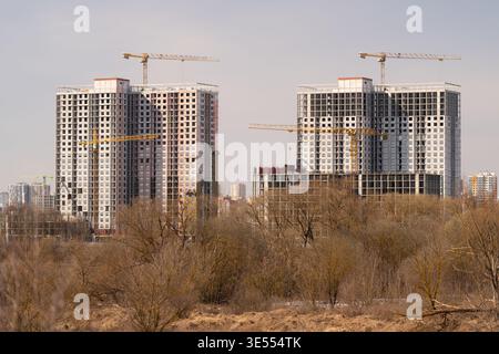 Bâtiments résidentiels en construction à proximité de broussailles, grues à tour ponctuant la skyline, dalles de béton apparentes, silhouettes de ville lointaine, Li d'automne Banque D'Images