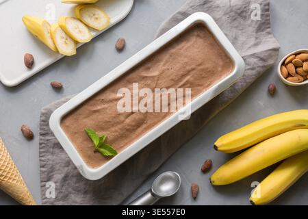 Crème glacée maison au chocolat à la banane dans un récipient métallique avec des grains de café et des amandes sur fond de béton gris. Espace pour le texte. Vue de dessus. Manger propre. Sans sucre. Banque D'Images