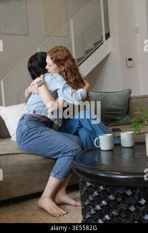 Deux femelles embrassant, assis sur le canapé en tissu à la maison près de la table basse, tenant des tasses à café. Embrasser, amis, confortable, intime, détendu, Lifestyle, Living || autorisation du modèle Banque D'Images
