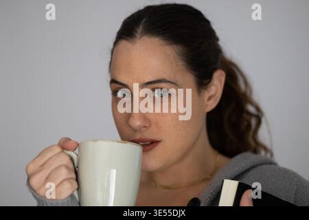 Femme tenant une tasse à café et un livre, se relaxant à l'intérieur sur un fond gris Banque D'Images