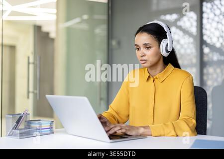 Femme concentrée portant des écouteurs blancs et une chemise jaune, assise à un bureau et tapant sur un ordinateur portable argenté, s'engageant dans le travail à distance ou la communication en ligne dans un environnement de bureau contemporain Banque D'Images