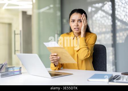 Femme d'affaires stressée ressentant le choc et le désespoir tout en lisant une lettre ou un avis désagréable dans une enveloppe au sujet de la perte d'emploi inattendue, la mise à pied, ou des problèmes financiers dans un bureau moderne Banque D'Images