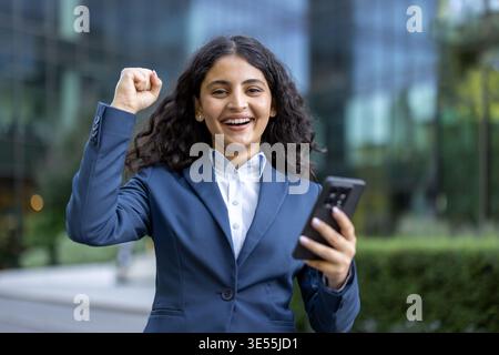 Jeune femme d'affaires aux cheveux bouclés foncés célébrant une réussite ou une victoire, exprimant l'excitation et la joie tout en regardant son smartphone dans un environnement d'entreprise urbain Banque D'Images