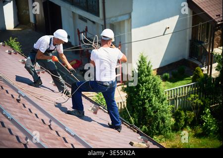 Hommes ouvriers installant un système de panneau solaire sur le toit de la maison. Électriciens en casques soulevant le module solaire photovoltaïque avec l'aide de cordes à l'extérieur. Concept d'énergie alternative et renouvelable. Banque D'Images