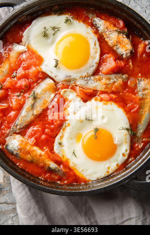 Petit déjeuner léger œufs frits avec sardines dans une sauce tomate épicée gros plan dans une poêle sur la table. Vue de dessus verticale Banque D'Images