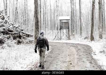 Un randonneur solitaire avec un sac à dos marche le long d'une route forestière enneigée vers un stand de chasse rustique élevé, véhiculant la solitude, l'exploration et l'hiver tranquille Banque D'Images