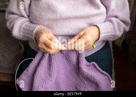 Femme âgée tricotant un pull violet à la main, gros plan des mains attentionnées et du fil Banque D'Images