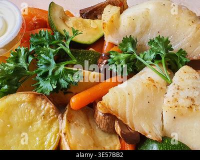 Vue rapprochée du ciel d'un repas préparé composé de filets de poisson blanc cuits au four et de légumes rôtis, dont des pommes de terre en tranches dorées et de l'orange Banque D'Images