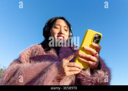 Femme asiatique dans un manteau de fourrure rose à l'aide d'un smartphone à l'extérieur. Elle porte des écouteurs, souriant tout en s'engageant avec du contenu numérique, incarnant moderne, urbain Banque D'Images