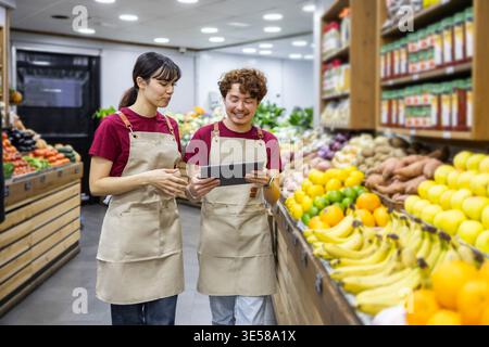 Deux vendeurs joyeux en uniforme se tiennent au milieu de la section des fruits vibrante d'une épicerie, discutant de l'inventaire sur une tablette numérique, entourent Banque D'Images