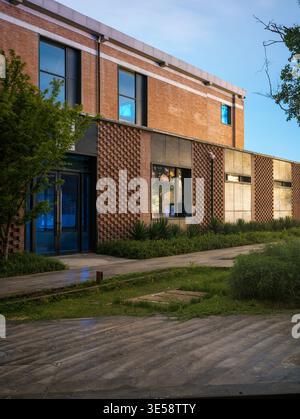 Bâtiment commercial rénové en brique rouge, aux fenêtres modernes teintées de bleu, donnant sur une terrasse pavée et en bois mouillé au crépuscule. La scène représente styl Banque D'Images