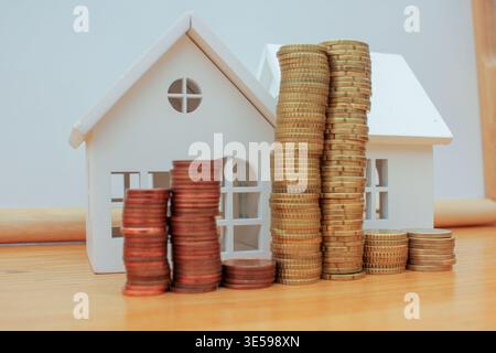 Image conceptuelle de petits modèles de maisons en bois placés derrière des tours de pièces de cents en euros, symbolisant la hausse des prix de l'immobilier, l'inflation, et le difficul Banque D'Images