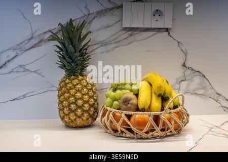 Gros plan de fruits frais, y compris l'ananas, les bananes et les raisins dans un panier sur un comptoir de cuisine moderne propre avec mur de marbre Banque D'Images