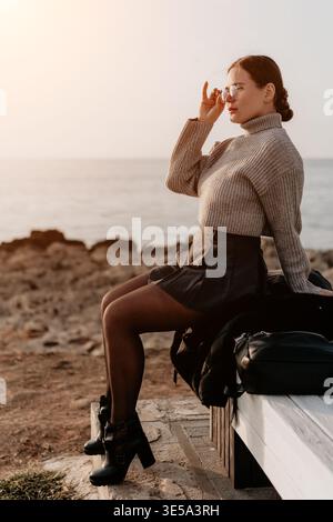 Femme coucher de soleil côte assis sur un banc surplombant l'océan tout en ajustant ses lunettes pendant une heure dorée sereine Banque D'Images