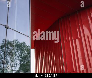 Gros plan sur un détail architectural montrant un contraste marqué entre une façade en verre réfléchissant et un panneau mural ondulé rouge vif. La scène représente Banque D'Images