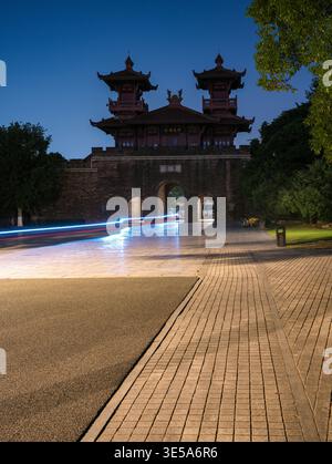 La porte d'entrée traditionnelle chinoise historique en brique illuminée contre un ciel nocturne sombre, avec des traînées de lumière rouge et blanche floues passant rapidement dans le Banque D'Images